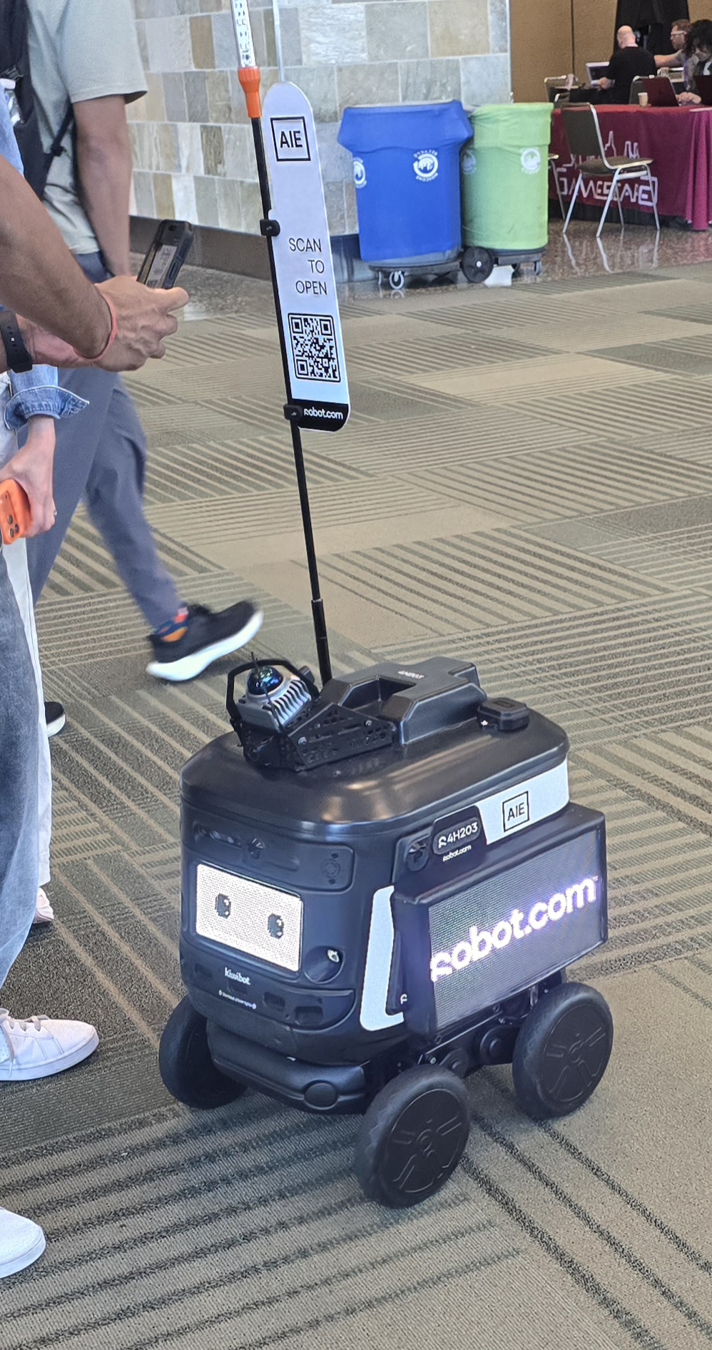 A Kiwibot delivery robot with a scan-to-open QR flag in the hallway